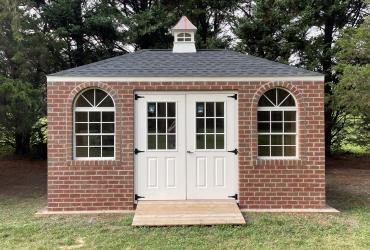 a brick hip roof shed. White double doors are accompanied by windows on either side and a cupola on top for added style.