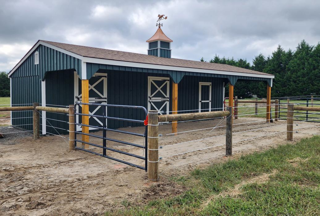 shed row barn with green/blue siding and complimentary wooden posts. fenced in space.