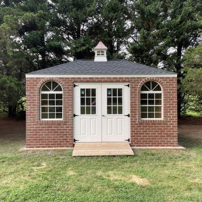 a brick hip roof shed. White double doors are accompanied by windows on either side and a cupola on top for added style.